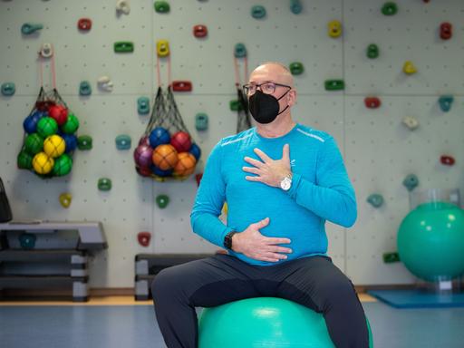 Ein Patient balanciert auf einem Sitzball und hat die Hände für eine Atemübung auf Brustkorb und Bauch gelegt.
