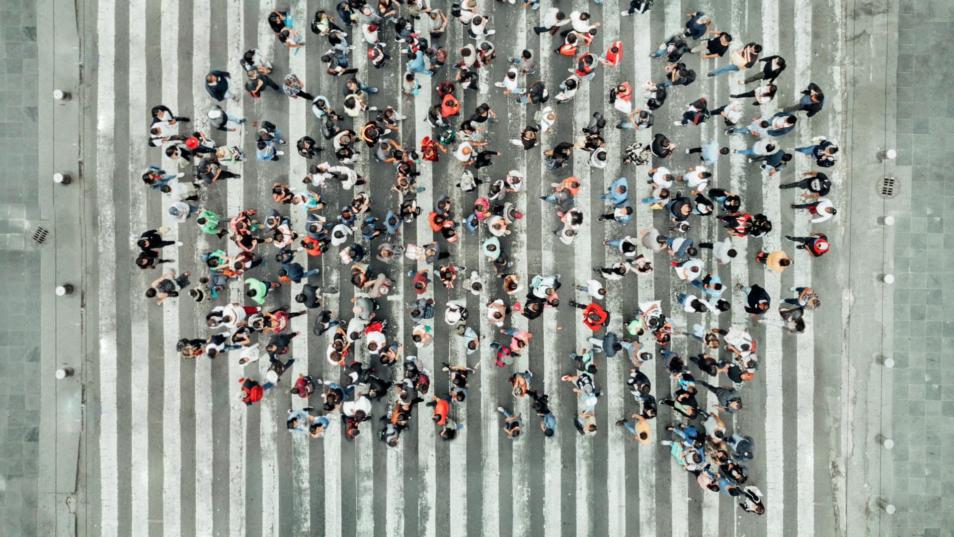 Vogelperspektive auf eine Gruppe von Menschen, die auf einem Zebrastreifen steht und gemeinsam die Form einer Sprechblase bildet.