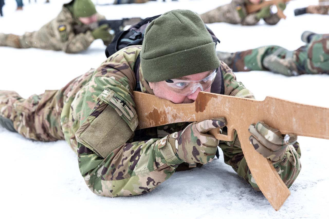 Ukrainische Zivilisten trainieren den Umgang mit der Waffe. Sie tragen Tarnanzüge und liegen im Schnee. Im Bildvordergrund hält ein Mann eine Waffenattrappe im Anschlag.