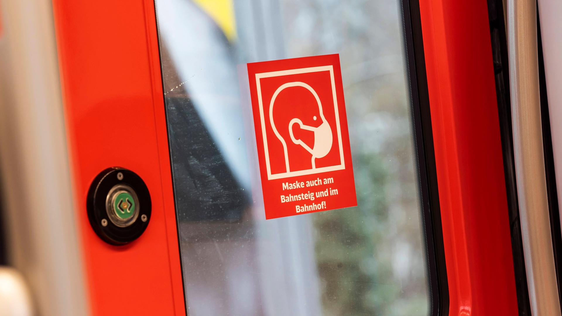 An der Tür einer Bahn klebt ein roter Aufkleber, auf dem eine Person mit Maske zu sehen ist. Darunter steht: "Maske auch am Bahnsteig und im Bahnhof!"