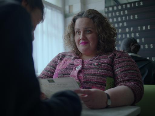 Szene aus der Serie "Rentierbaby": Eine lächelnde Frau (Jessica Gunning) sitzt an einem Schreibtisch einer anderen Person gegenüber und hält einen Brief in der Hand. 