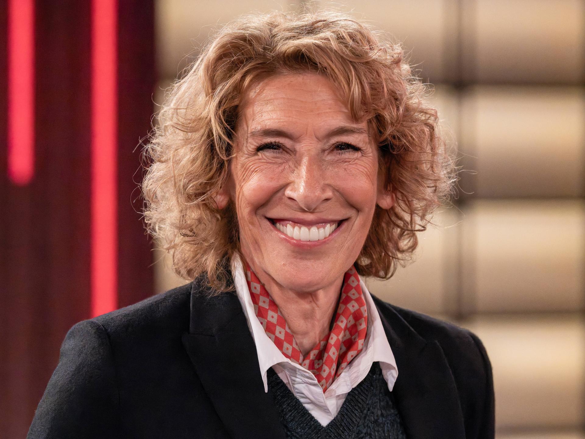 Die österreichische Schauspielerin Adele Neuhauser lacht in die Kamera. Sie trägt ein schwarzes Oberteil, darunter eine weiße Bluse und ein rot gemustertes Halstuch.