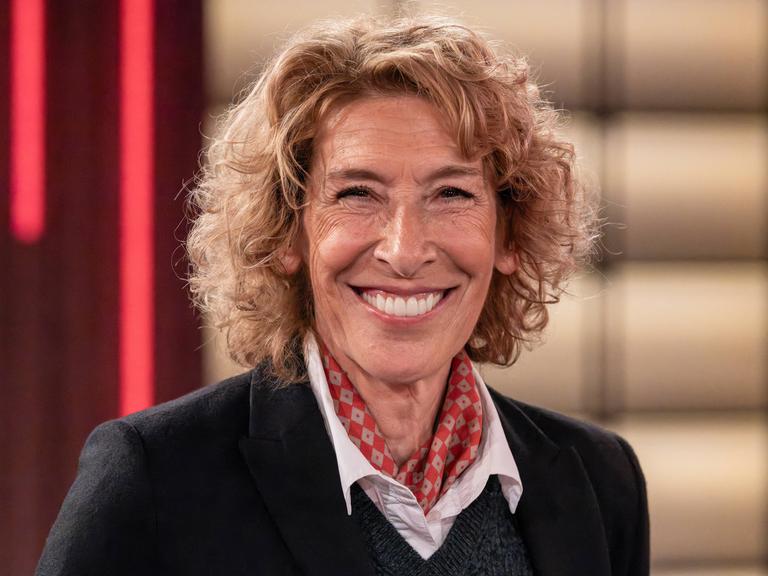 Die österreichische Schauspielerin Adele Neuhauser lacht in die Kamera. Sie trägt ein schwarzes Oberteil, darunter eine weiße Bluse und ein rot gemustertes Halstuch.