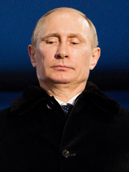 Portrait von Putin, der einen dunklen Mantel trägt, vor einer blauen Wand steht und nach unten schaut.