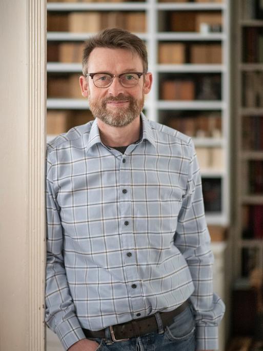 Christoph Peters steht an einen Türrahmen gelehnt, im Hintergrund ist ein Bücherregal zu sehen.