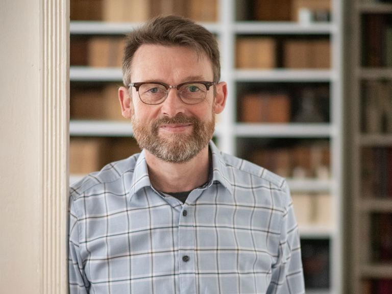 Christoph Peters steht an einen Türrahmen gelehnt, im Hintergrund ist ein Bücherregal zu sehen.