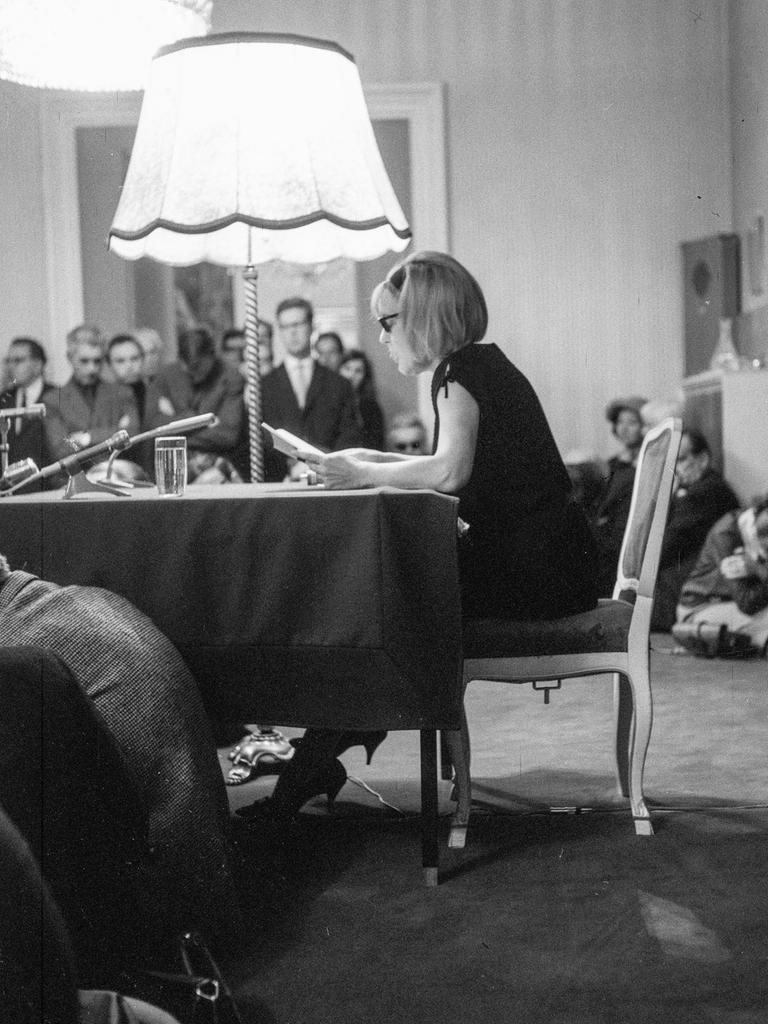 Die Schriftstellerin Ingeborg Bachmann sitzt bei einer Lesung auf einem altmodisch wirkenden Stuhl, neben ihr eine große Stehlampe. Auf dem Tisch vor ihr einige Mikrofone.