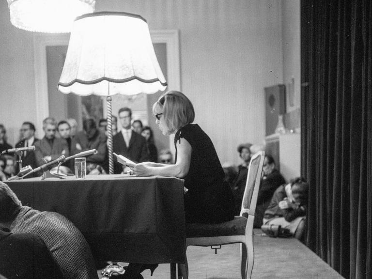 Die Schriftstellerin Ingeborg Bachmann sitzt bei einer Lesung auf einem altmodisch wirkenden Stuhl, neben ihr eine große Stehlampe. Auf dem Tisch vor ihr einige Mikrofone.