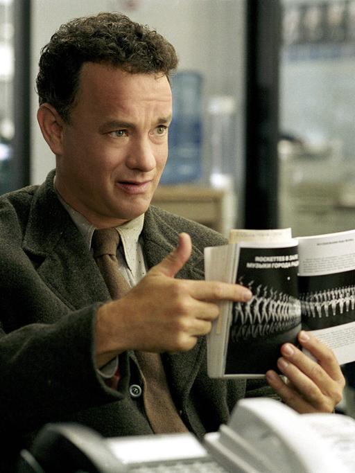 Der Schauspieler Tom Hanks zeigt im Film "Terminal" mit dem Finger auf die Seite eines Buches, die ein Foto einer Reihe von Tänzerinnen zeigt. Im Hintergrund idt ein Computer zu sehen.