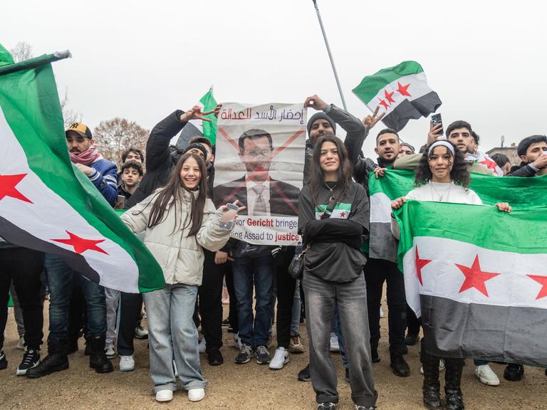 Syrerinnen und Syrer feiern den Fall des Assad-Regimes, sie tragen syrische Flaggen und ein durchgestrichenes Porträt von Assad.