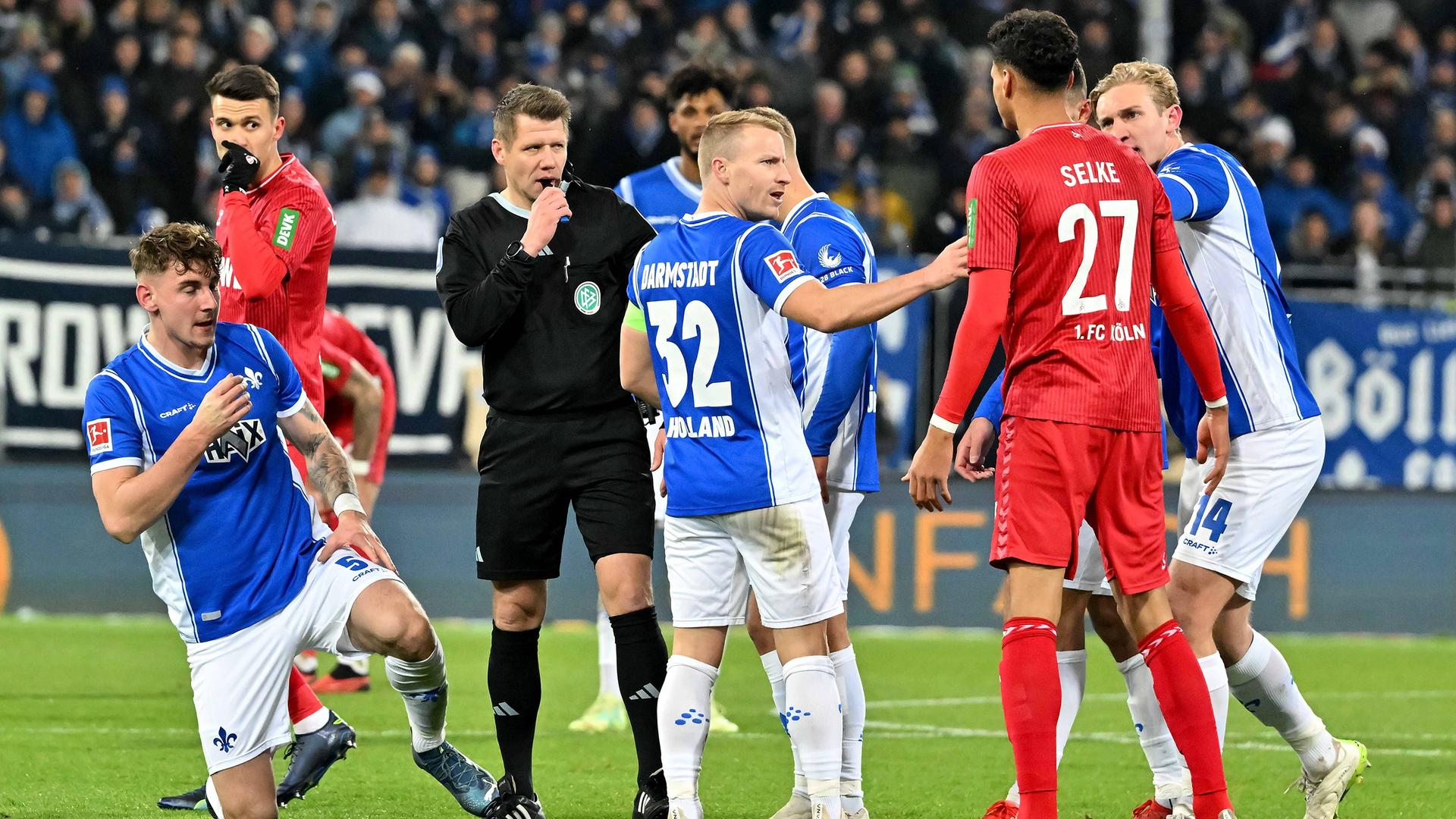Szene aus dem Bundesliga-Spiel Darmstadt gegen Köln; der Schiedsrichter pfeift, um ihn herum stehen Spieler beider Mannschaften.