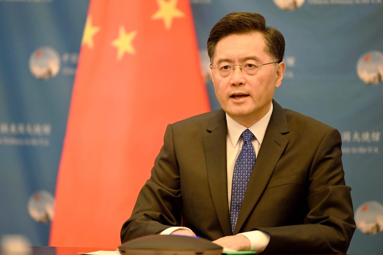 Ein Chinese mit randloser Brille und Anzug sitzt an einem Tisch und spricht. Im Hintergrund ist die chinesische Flagge zu sehen.