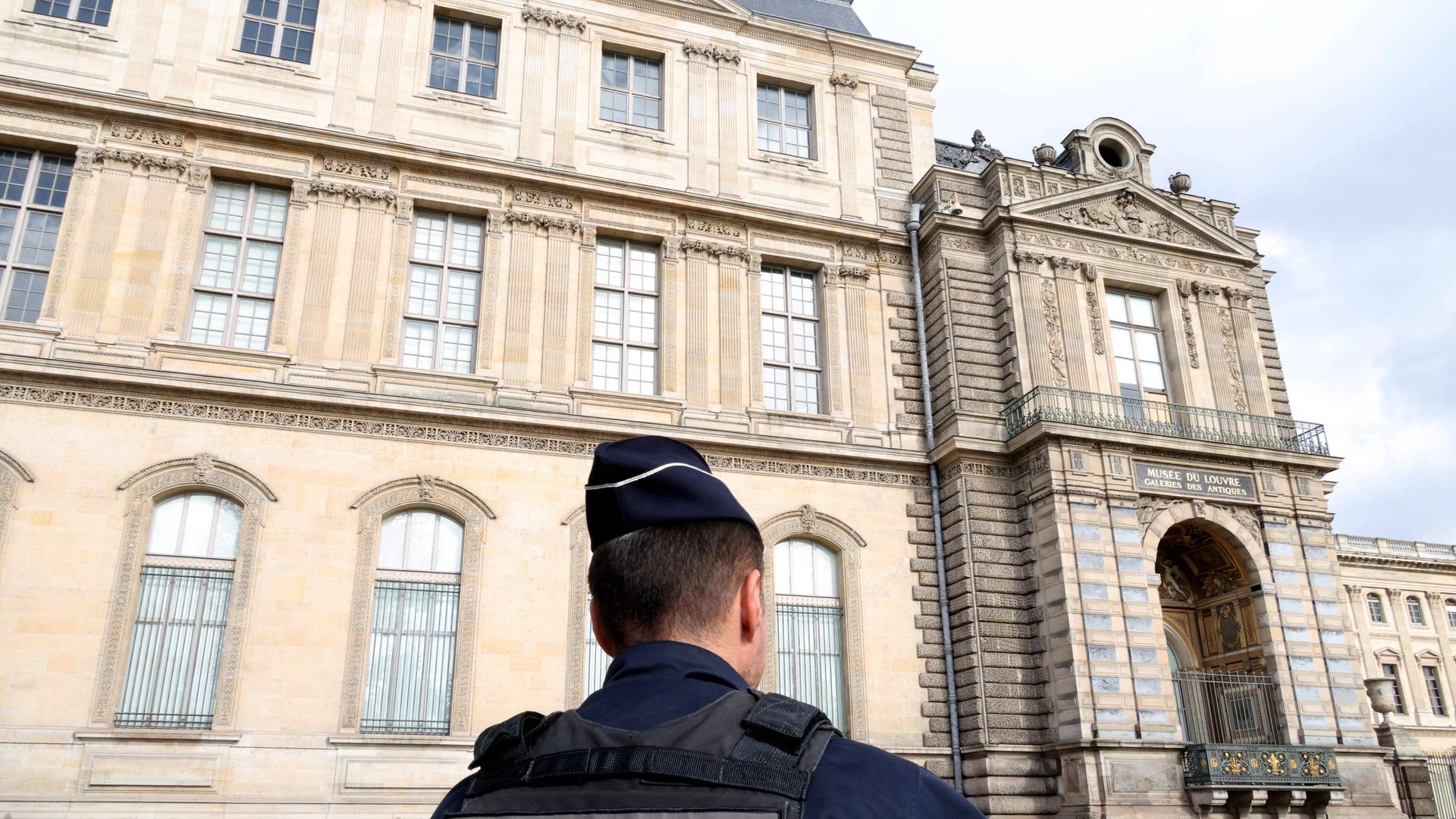 Vor dem Gebäudeteil steht ein schwarz gekleideter Polizist. Man sieht ihn von hinten.