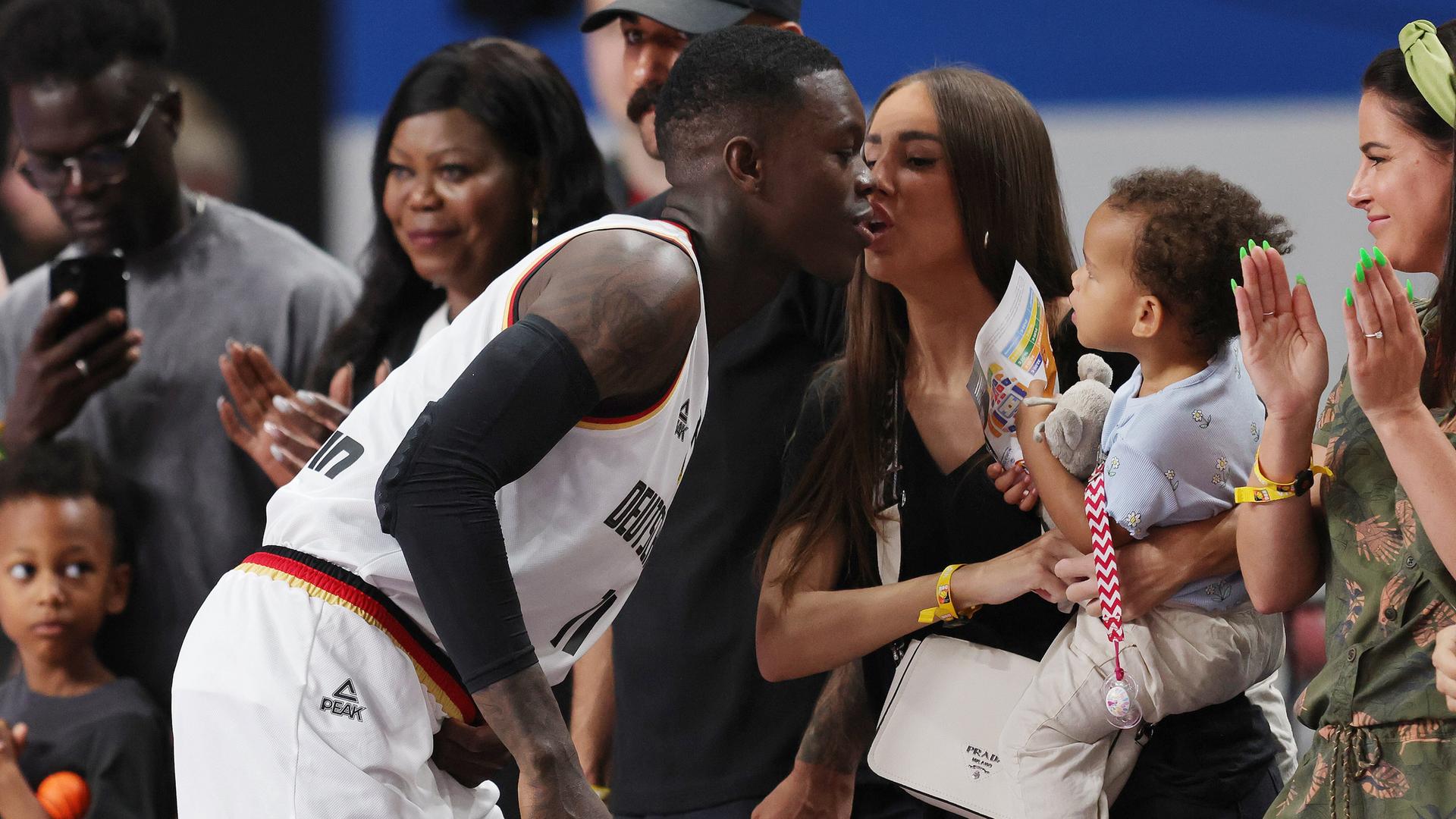 Das Foto zeigt Basketball-Nationalspieler Dennis Schröder. Er beugt sich zu seiner Frau Ellen und ist in Begriff, ihr einen Kuss zu geben. Ellen Schröder hat eines der Kinder auf ihrem Arm, um die drei herum stehen weitere Menschen. Das Foto zeigt Basketball-Nationalspieler Dennis Schröder. Er beugt sich zu seiner Frau Ellen und ist in Begriff, ihr einen Kuss zu geben. Ellen Schröder hat eines der Kinder auf ihrem Arm, um die drei herum stehen weitere Menschen.