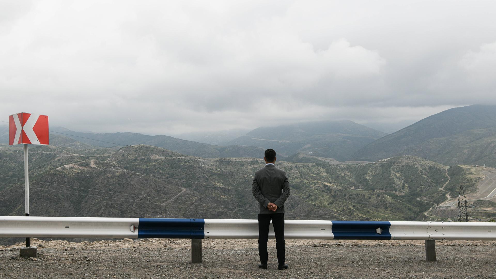 Ein Mann steht mit dem Rücken zur Kamera an einer Leitplanke und blickt auf ein Bergpanorama.