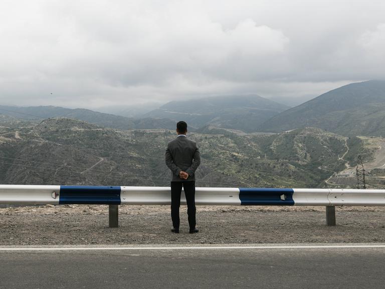 Ein Mann steht mit dem Rücken zur Kamera an einer Leitplanke und blickt auf ein Bergpanorama.