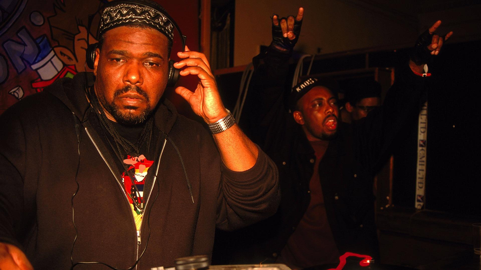 Afrika Bambaataa steht mit Kopfhörern an einem DJ-Pult.