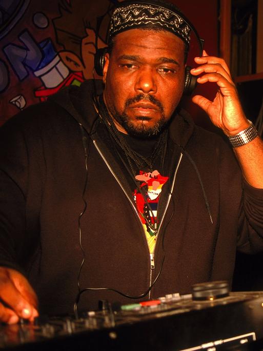 Afrika Bambaataa steht mit Kopfhörern an einem DJ-Pult.