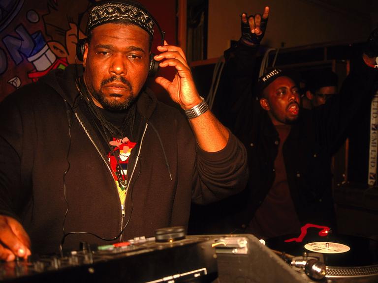 Afrika Bambaataa steht mit Kopfhörern an einem DJ-Pult.
