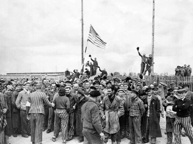 Menschen drängen sich auf einem Hof und stehen auf Zäunen, die meisten von ihnen in gestreifter Häftlingskleidung, winken und schwenken eine amerikanische Flagge.
