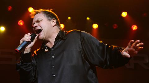 Ein Mann steht mit ausgebreiteten Armen auf einer Bühne und singt. In der rechten Hand hält er ein Mikrofon. Es ist der US-Rockmusiker Meat Loaf.