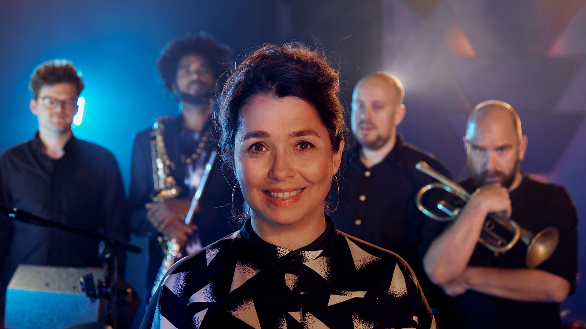 Sängerin Céline Rudoph steht auf einer beleuchteten Bühne mit ihren vier männlichen Bandmitgliedern, die teilweise ihre Blechblasinstrumente dabei haben und alle gemeinsam in die Kamera schauen.