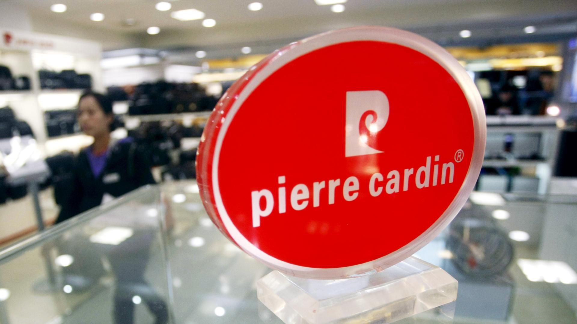 Das Logo von Pierre Cardin mit weißer Schrift in einem roten Oval steht auf einer gläsernen Verkaufsplatte. Das Logo von Pierre Cardin mit weißer Schrift in einem roten Oval steht auf einer gläsernen Verkaufsplatte.