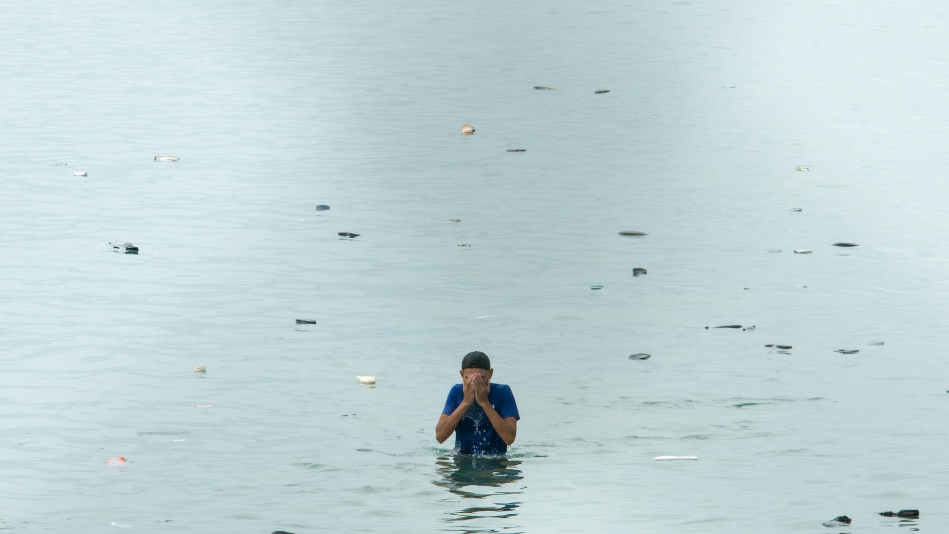 Ein Mensch steht im Wasser mit vorgehaltenen Händen, umringt von Schuhen. Ein Mensch steht im Wasser mit vorgehaltenen Händen, umringt von Schuhen.