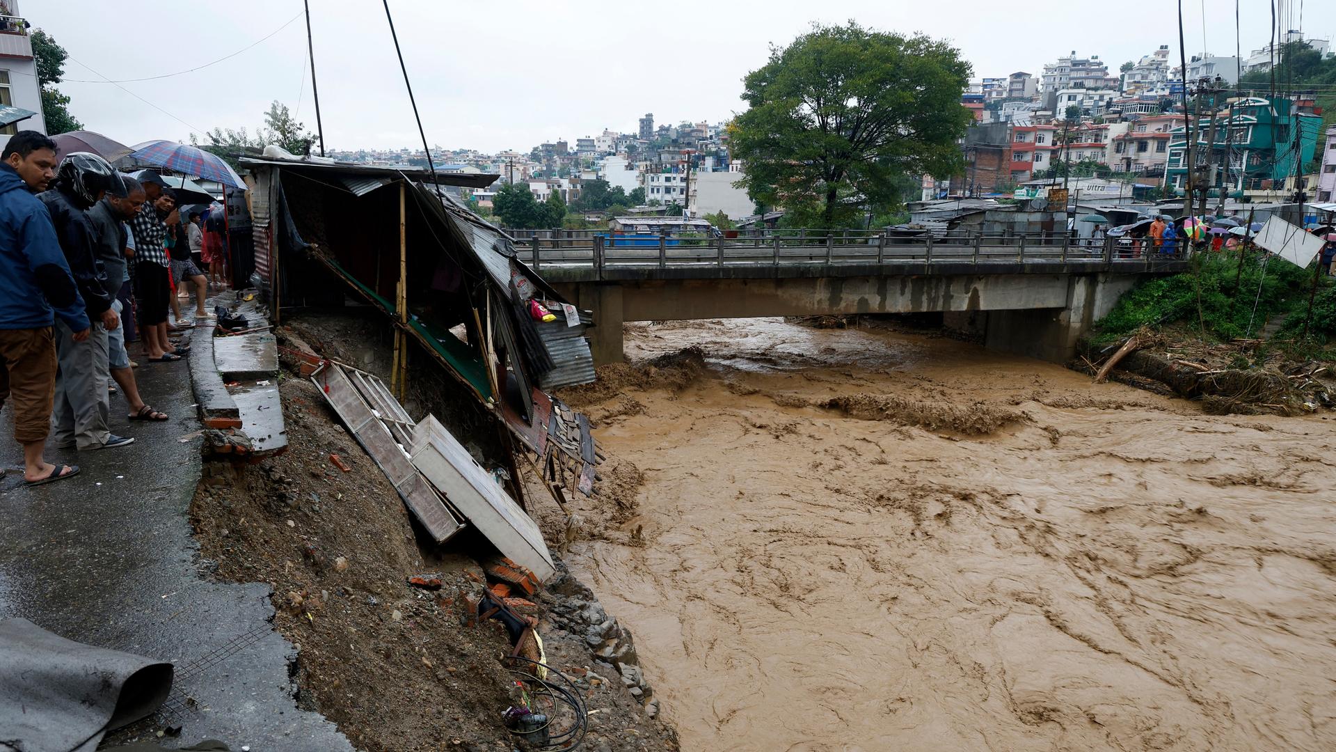 Szene aus Kathmandu in Nepal: Menschen mit Regenschirmen stehen neben Verwüstungen durch heftige Niederschläge am Ufer des Flusses Bagmati nach heftigen Regenfällen. Aufnahme vom 28.09.2024.