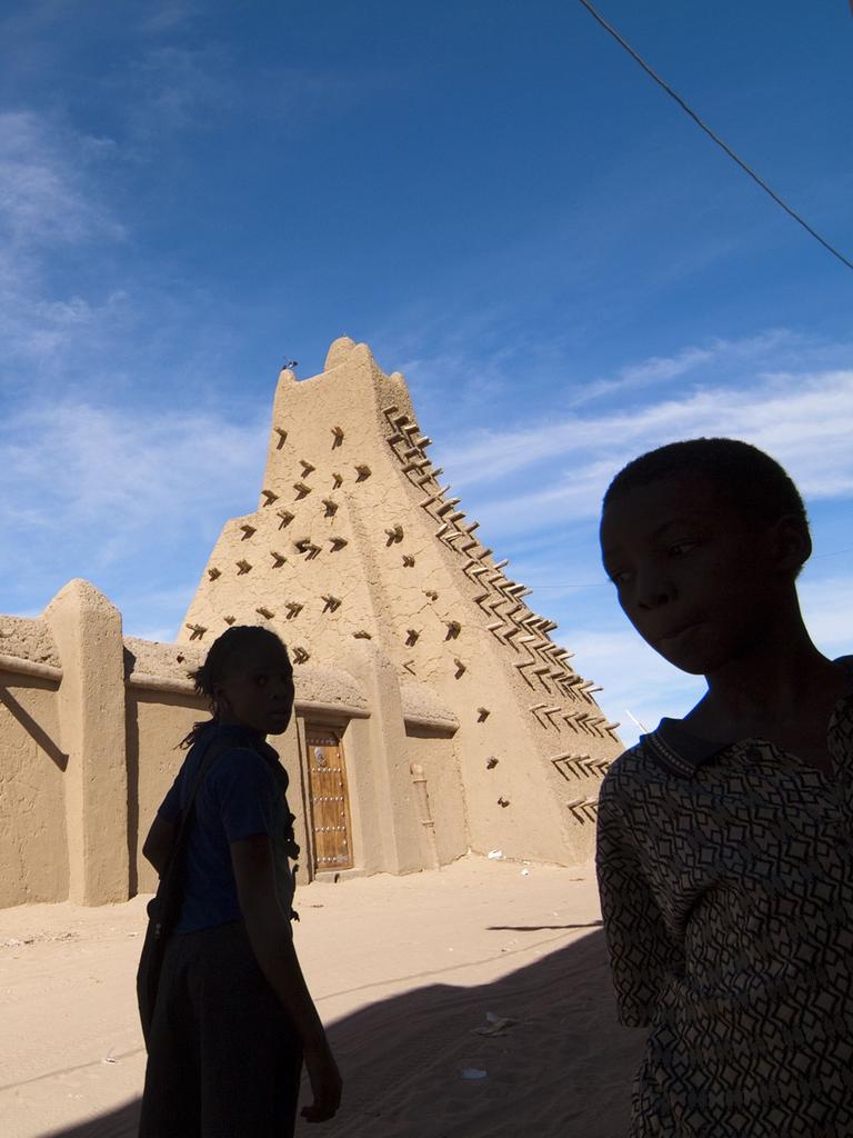 Blick auf die in Lehm gebaute Moschee vor blauem Himmel. Davor die Silhoutte zweier Kinder.