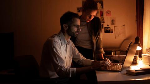 Die Schauspieler Hadi Khanjanpour als Khalil und Leyla Kristin Suckow als Leyla stehen in einem rötlich beschienenen Raum über einen Bildschirm gebeugt.