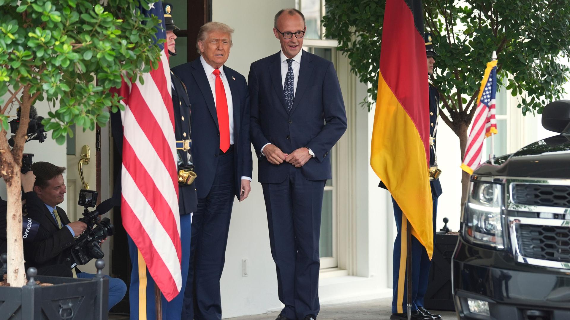 Bundeskanzler Merz und US-Präsident Trump stehen nebeneinander vor dem Weißen Haus. Neben ihnen hängen jeweils die Landesfahnen.
