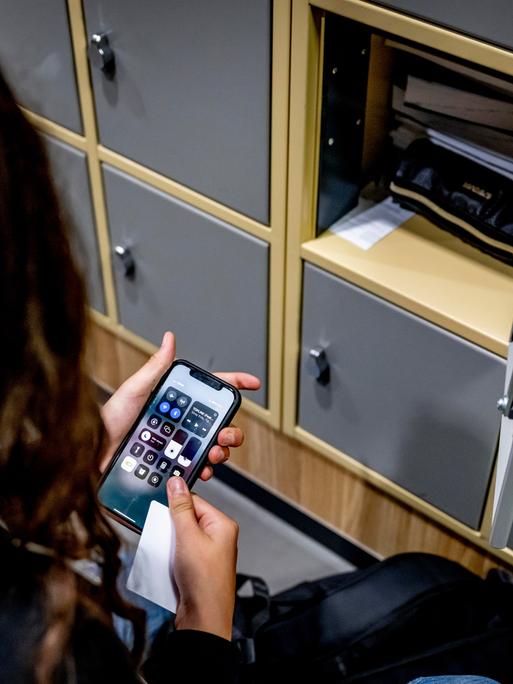 Eine Schülerin sitzt neben einem offenen Spind und hält ein Smartphone in der Hand