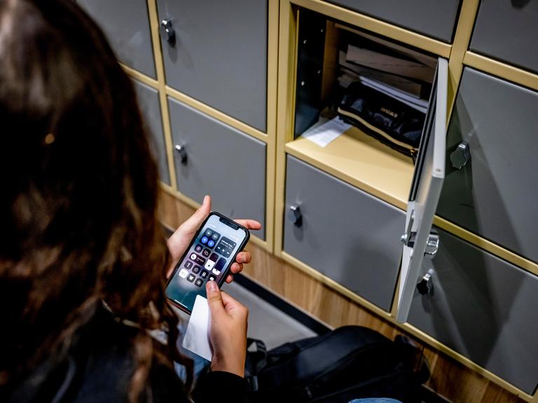 Eine Schülerin sitzt neben einem offenen Spind und hält ein Smartphone in der Hand
