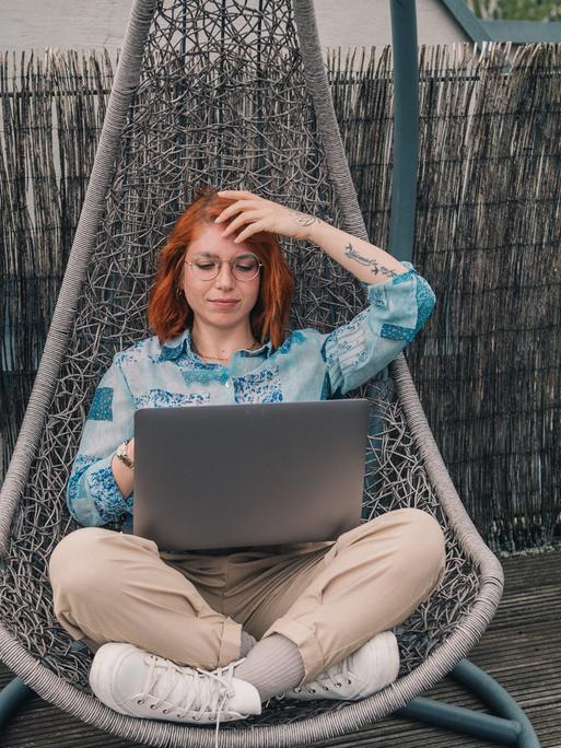 Eine Frau mit roten Haaren sitzt in einem Hängesessel auf dem Balkon und arbeitet am Laptop.
