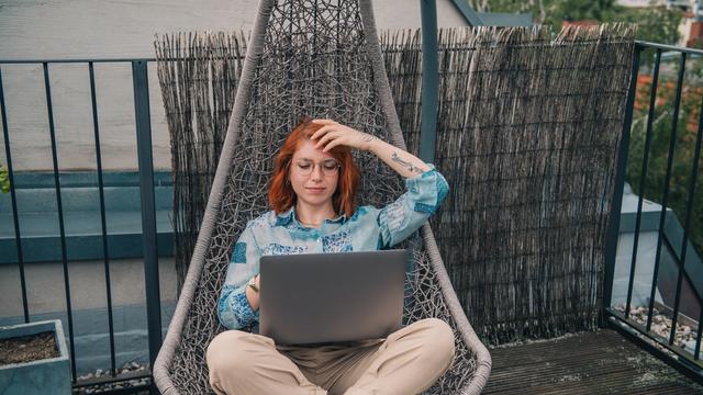 Eine Frau mit roten Haaren sitzt in einem Hängesessel auf dem Balkon und arbeitet am Laptop.