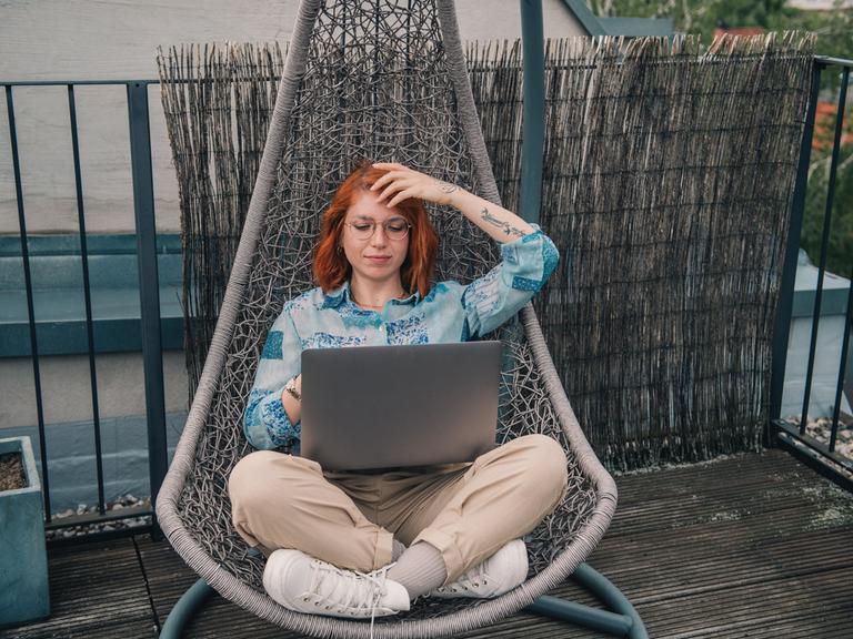 Eine Frau mit roten Haaren sitzt in einem Hängesessel auf dem Balkon und arbeitet am Laptop.