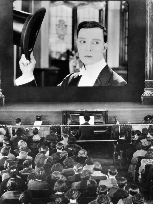 Schwarz-Weiß-Foto eines voll besetzten Kinos in New York im Jahr 1925, das einen Stummfilm mit Buster Keaton zeigt. Dazu spielt ein Klavierspieler.