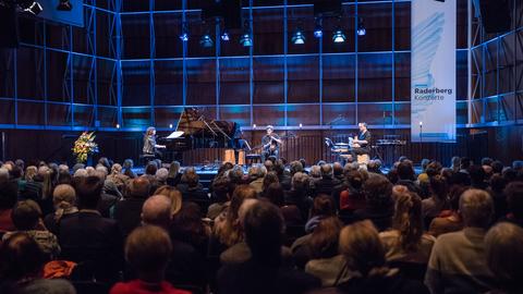 Man sieht eine Bühne, auf der drei Musiker ein Konzert geben. 