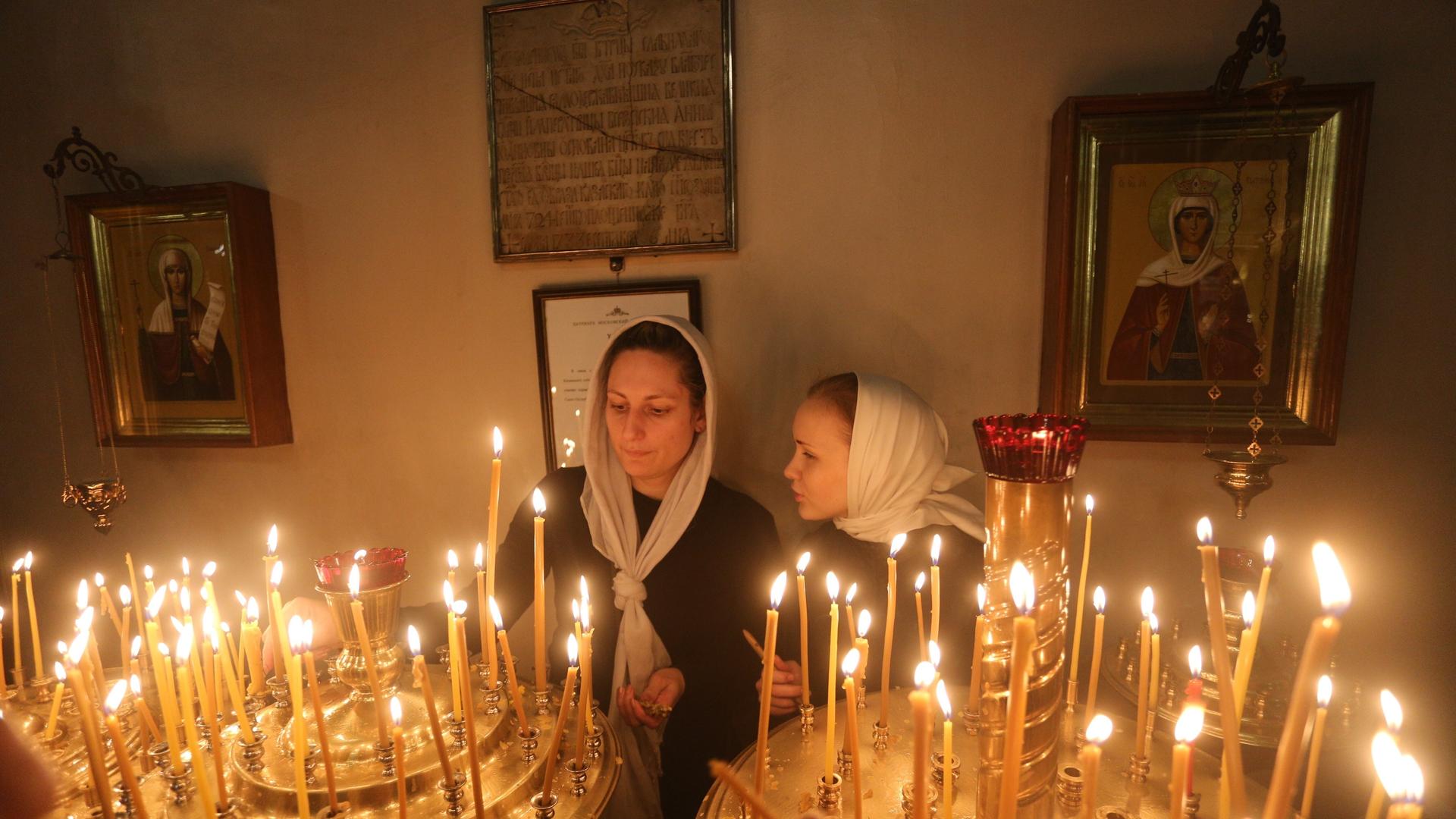 Zwei Frauen mit je einem Tuch über dem Kopf zünden während eines orthodoxen Gottesdienstes in der Kazaner Kathedrale in St. Petersburg Kerzen an, hinter ihnen sind Ikonen zu sehen.