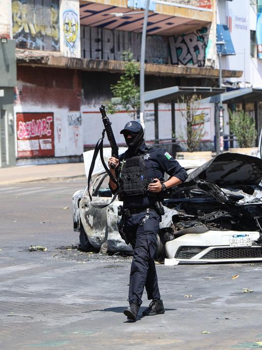 Ein Polizist läuft neben einem ausgebrannten Auto auf einer Straße entlang. Er hält ein Gewehr in der Hand.