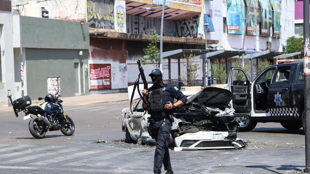 Ein Polizist läuft neben einem ausgebrannten Auto auf einer Straße entlang. Er hält ein Gewehr in der Hand.
