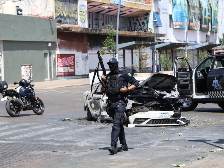 Ein Polizist läuft neben einem ausgebrannten Auto auf einer Straße entlang. Er hält ein Gewehr in der Hand.
