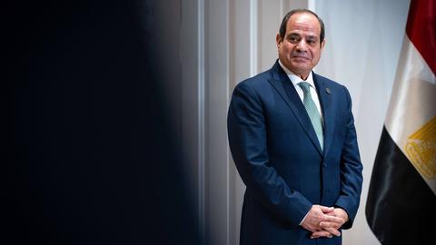 Der ägyptische Präsident Abdel Fattah El-Sisi steht neben der ägyptischen Flagge und schaut links aus dem Bild heraus. Er trägt einen blauen Anzug und hat die Hände vor dem Bauch gefalltet.