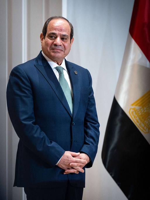 Der ägyptische Präsident Abdel Fattah El-Sisi steht neben der ägyptischen Flagge und schaut links aus dem Bild heraus. Er trägt einen blauen Anzug und hat die Hände vor dem Bauch gefalltet.