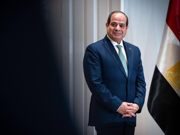 Der ägyptische Präsident Abdel Fattah El-Sisi steht neben der ägyptischen Flagge und schaut links aus dem Bild heraus. Er trägt einen blauen Anzug und hat die Hände vor dem Bauch gefalltet.