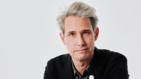 Ein Mann mit grauem, vollem Haar blickt nachdenklich in die Kamera. Er trägt ein graues Hemd unter einem schwarzen Jackett sowie einen roten Schottenrock. Er sitzt auf einem Stuhl.