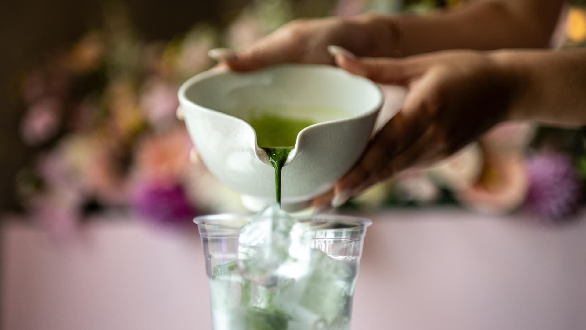 Eine Nahaufnahme zeigt, wie frisch gebrühter Matcha aus einer Keramikschale in einen Plastikbecher auf Eis gegossen wird.