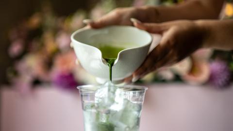 Eine Nahaufnahme zeigt, wie frisch gebrühter Matcha aus einer Keramikschale in einen Plastikbecher auf Eis gegossen wird.