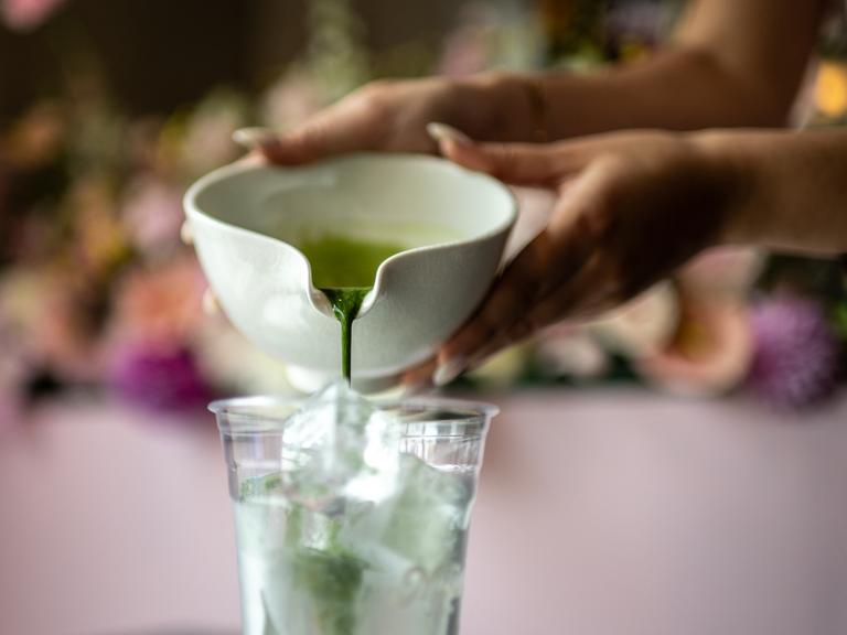 Eine Nahaufnahme zeigt, wie frisch gebrühter Matcha aus einer Keramikschale in einen Plastikbecher auf Eis gegossen wird.